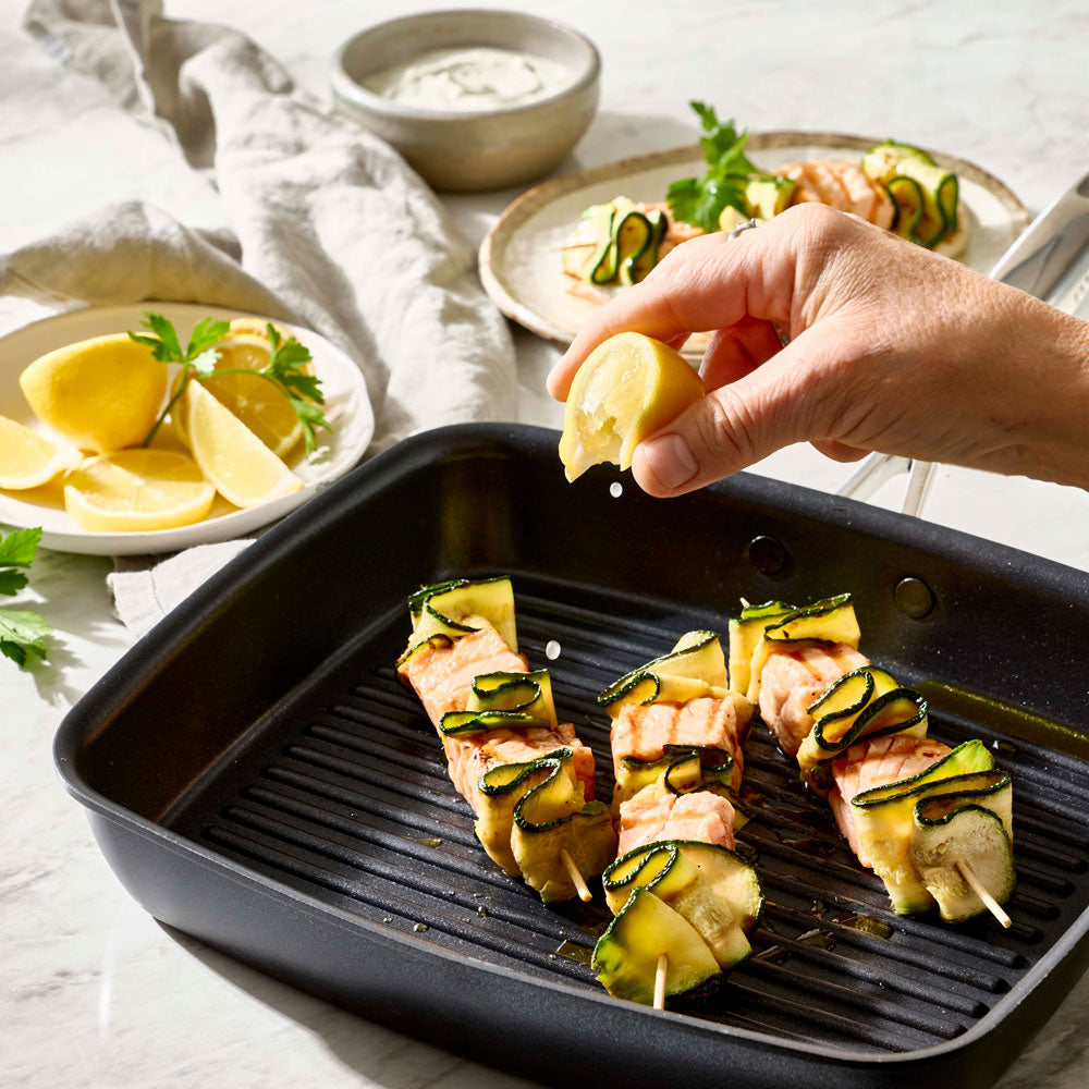 A hand is squeezing a lemon over food skewered in a black grill pan.