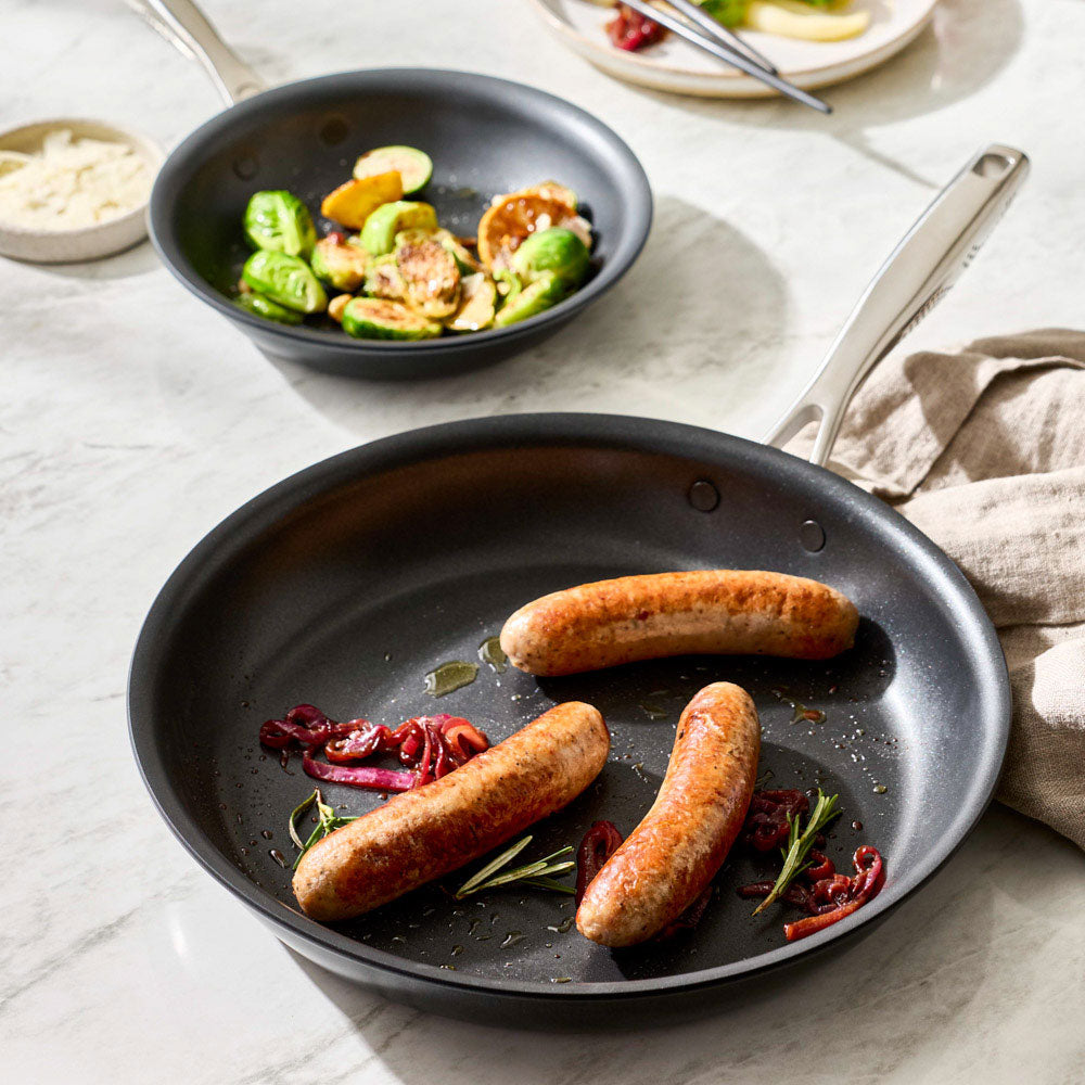 Sausages are cooking inside a black frying pan on a kitchen surface with another dish nearby.