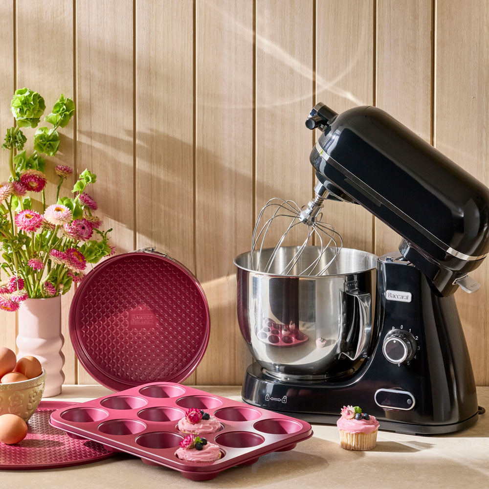 A black stand mixer with a stainless steel bowl sits on a countertop beside a pink muffin tin and a matching pink lid, with cupcakes and flowers in the background against a wood-paneled wall.