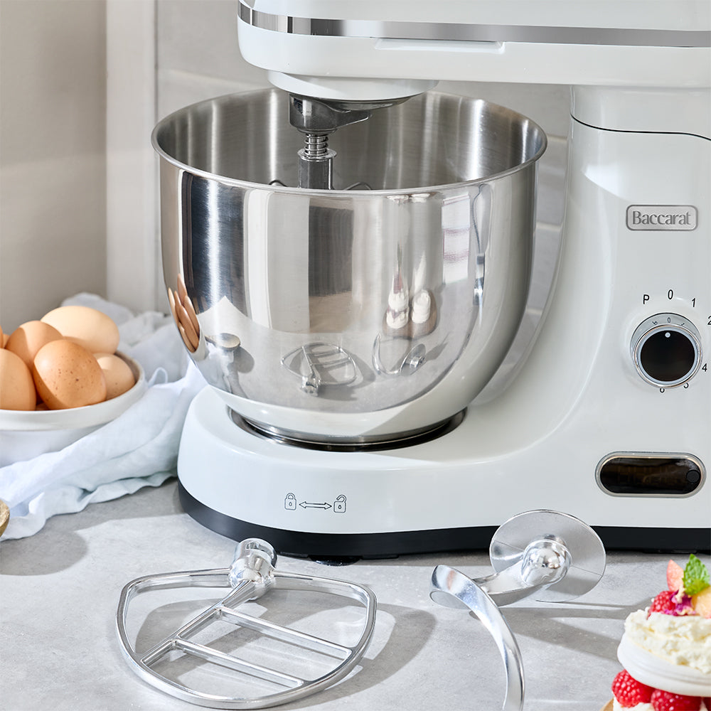White stand mixer with a silver bowl on a kitchen counter, surrounded by eggs and a cake.