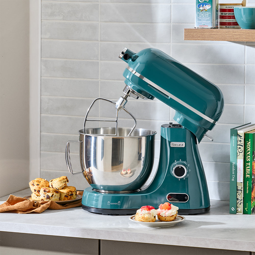 Teal stand mixer on a kitchen counter with baked goods and cookbooks in the background.