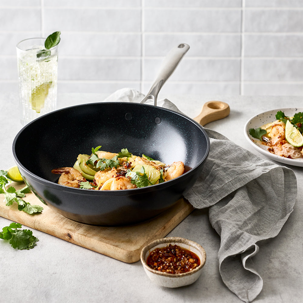The black frying pan is placed on a wooden board on a counter, filled with food, next to a cloth and a drink.