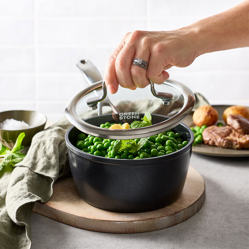 A person is lifting the glass lid off the black saucepan, which is filled with green vegetables.