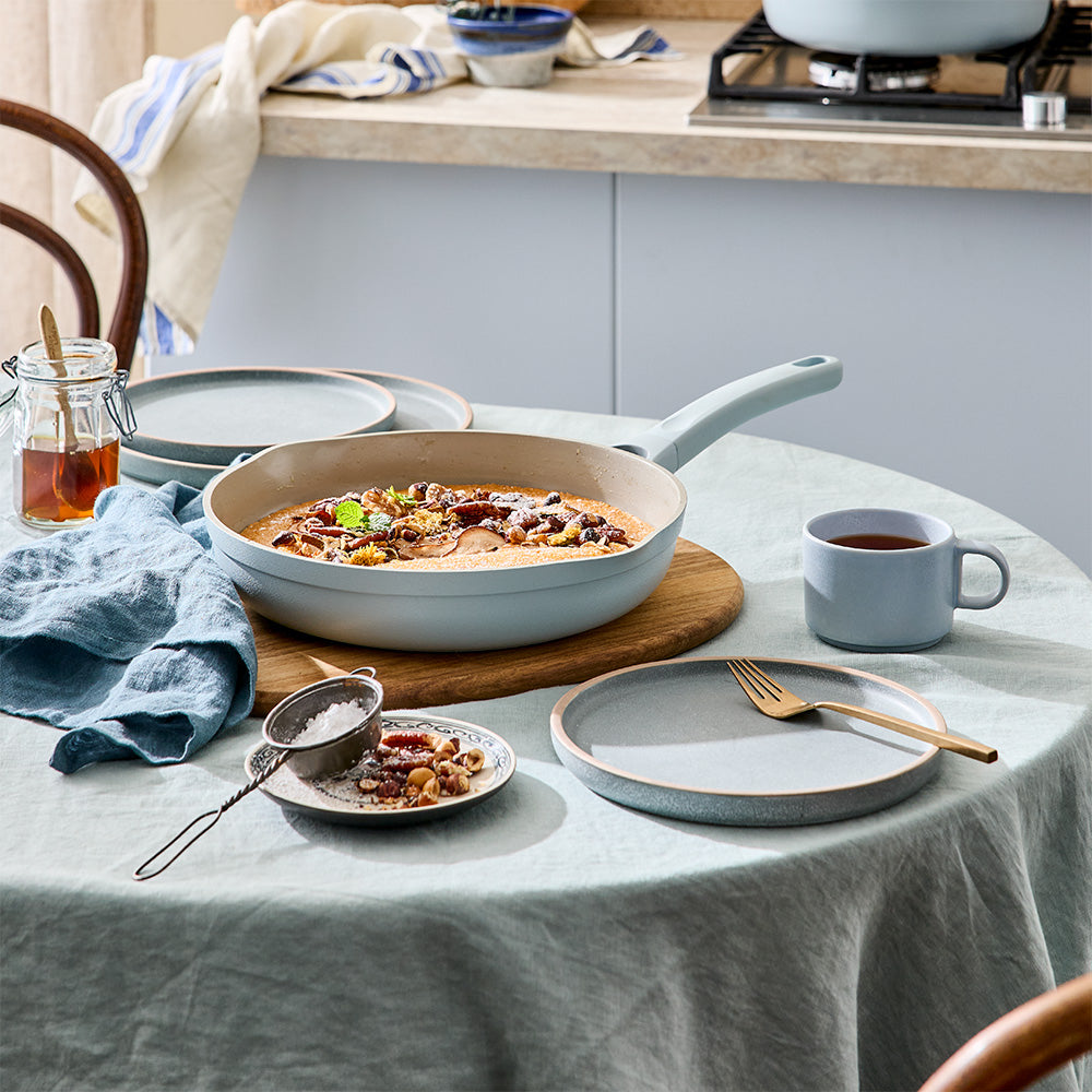 A dining table is set with plates, cutlery, tea cups, and a light blue frying pan containing food.