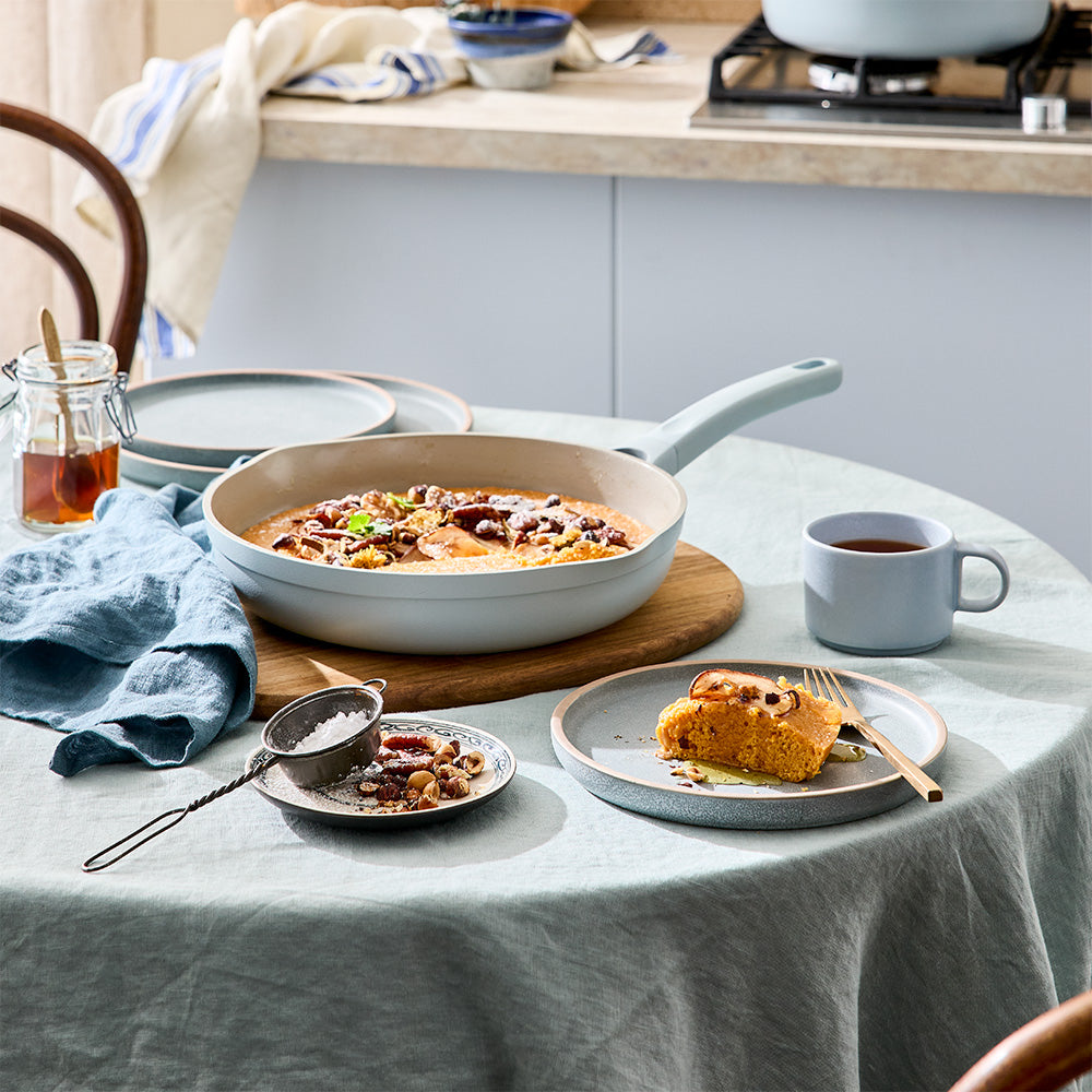 A light blue frying pan is set on a dining table next to served plates, cutlery, and tea cups.