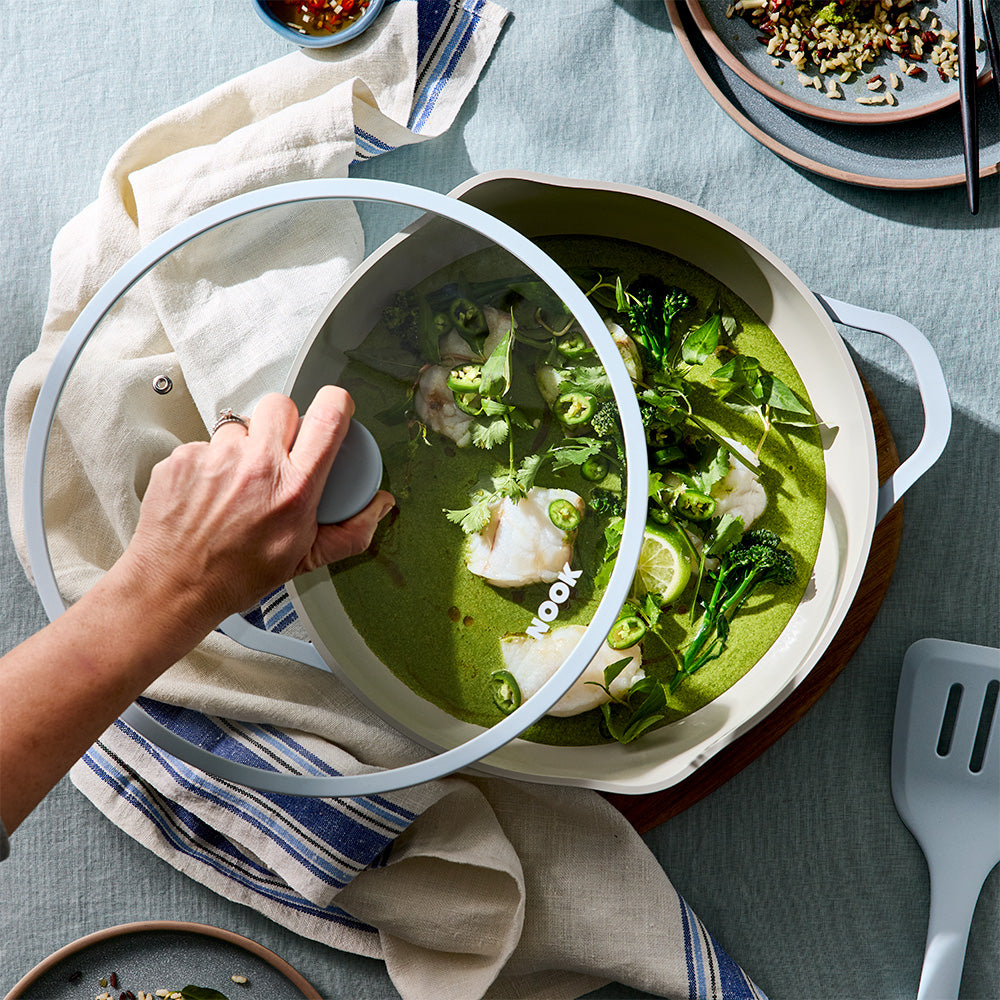A person's hand lifts the glass lid of a beige nonstick Dutch oven containing a green fish curry soup, set on a blue tablecloth with dining props.
