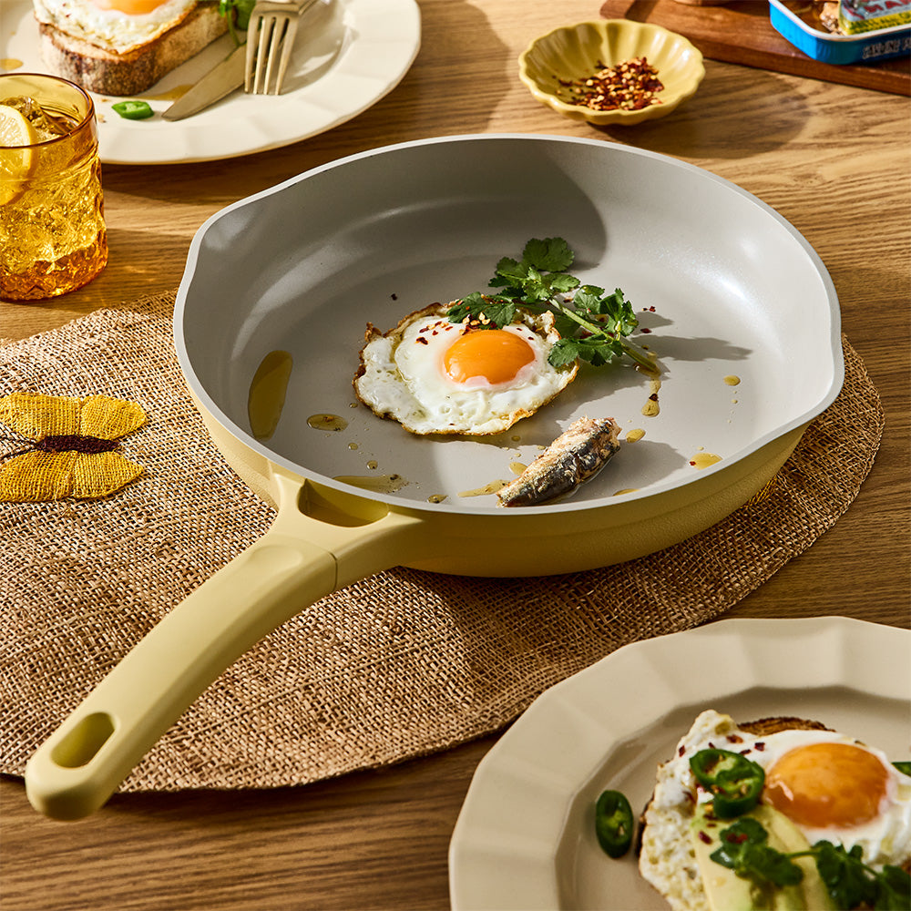 A light yellow non-stick frying pan with a matching handle, shown on a wooden table with breakfast food. The pan contains a fried egg with a bright orange yolk and green parsley garnish.