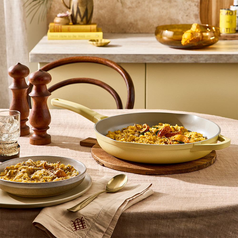 A light yellow enameled cast iron skillet filled with a rice dish, styled on a beige tablecloth in a warm, rustic dining setting with a matching bowl, wooden tableware, and salt and pepper shakers.