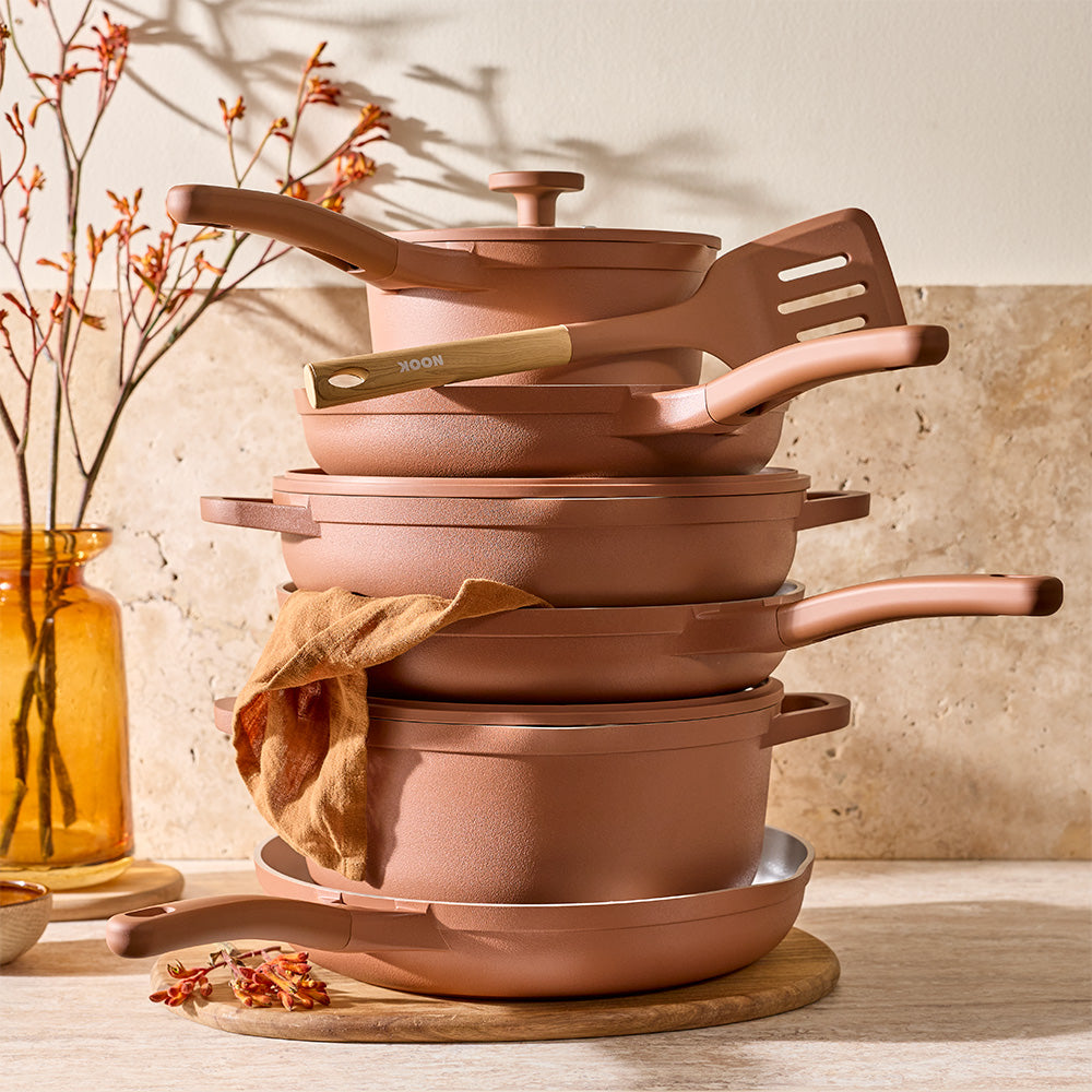 A set of nonstick cookware in a warm terracotta or dusty rose metallic finish, arranged on a wooden board on a tabletop with dried branches and an amber glass vase in the background.