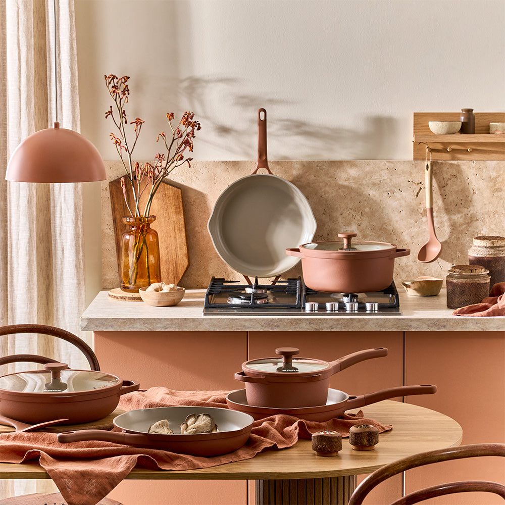 A wide shot shows a kitchen scene featuring blush pink cookware sets and wooden utensils.