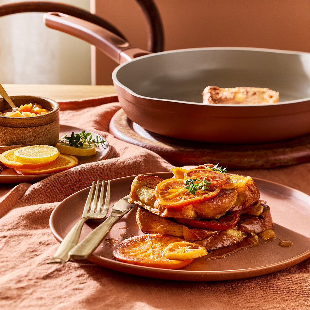 A close-up view shows cooked food being served from a terracotta-colored frying pan onto a plate next to citrus garnishes.