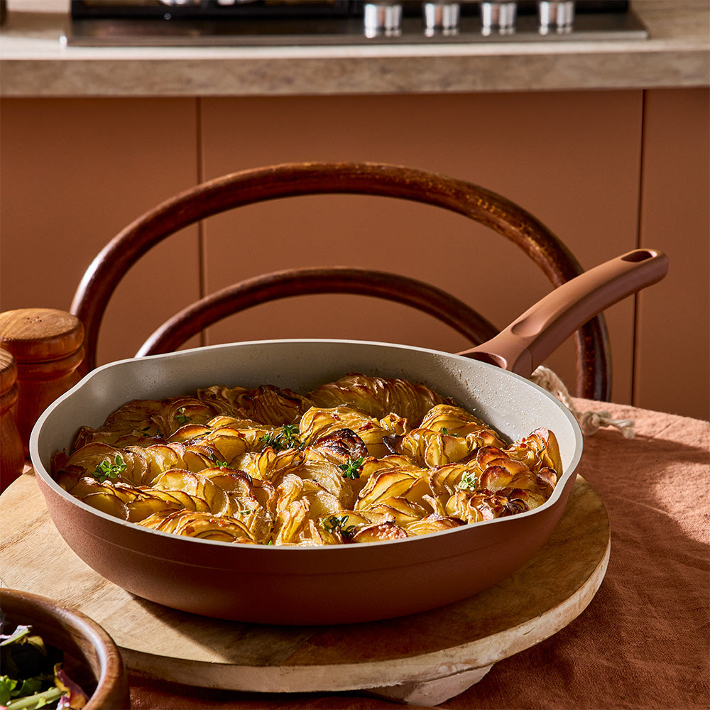 A brown non-stick frying pan with a light gray interior, filled with roasted artichokes, set on a wooden board in a warm kitchen environment.