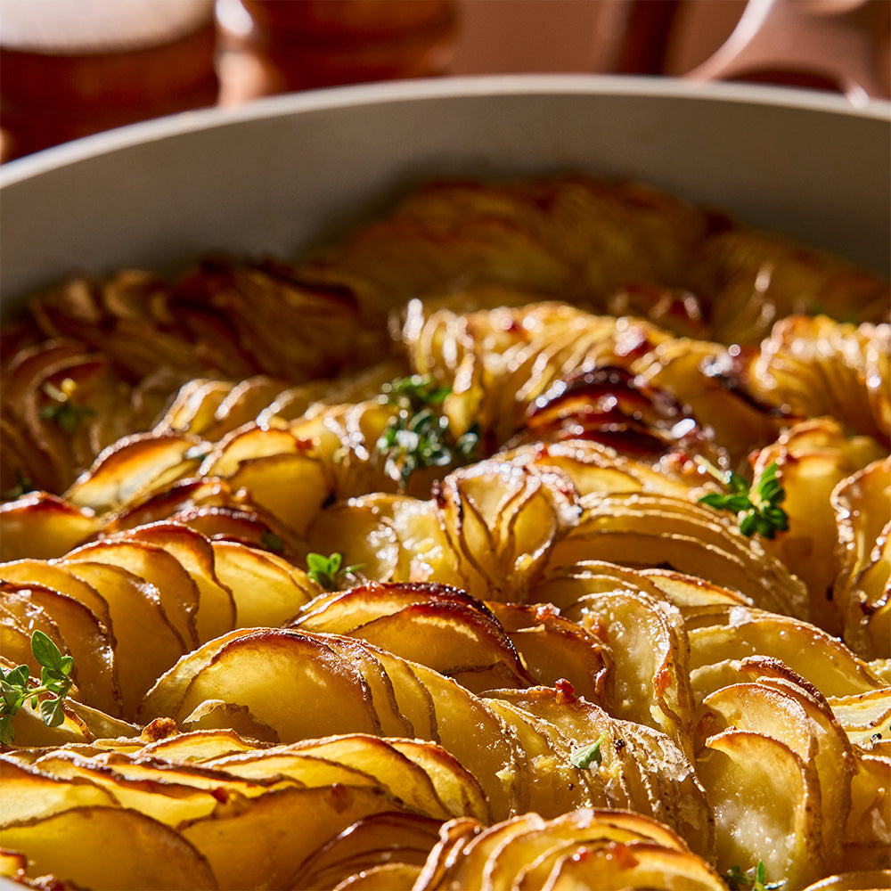 Close-up of a baked potato casserole or baked potato dish in a light-colored baking pan, with golden-brown potato slices, melted cheese, and small green herb garnishes.