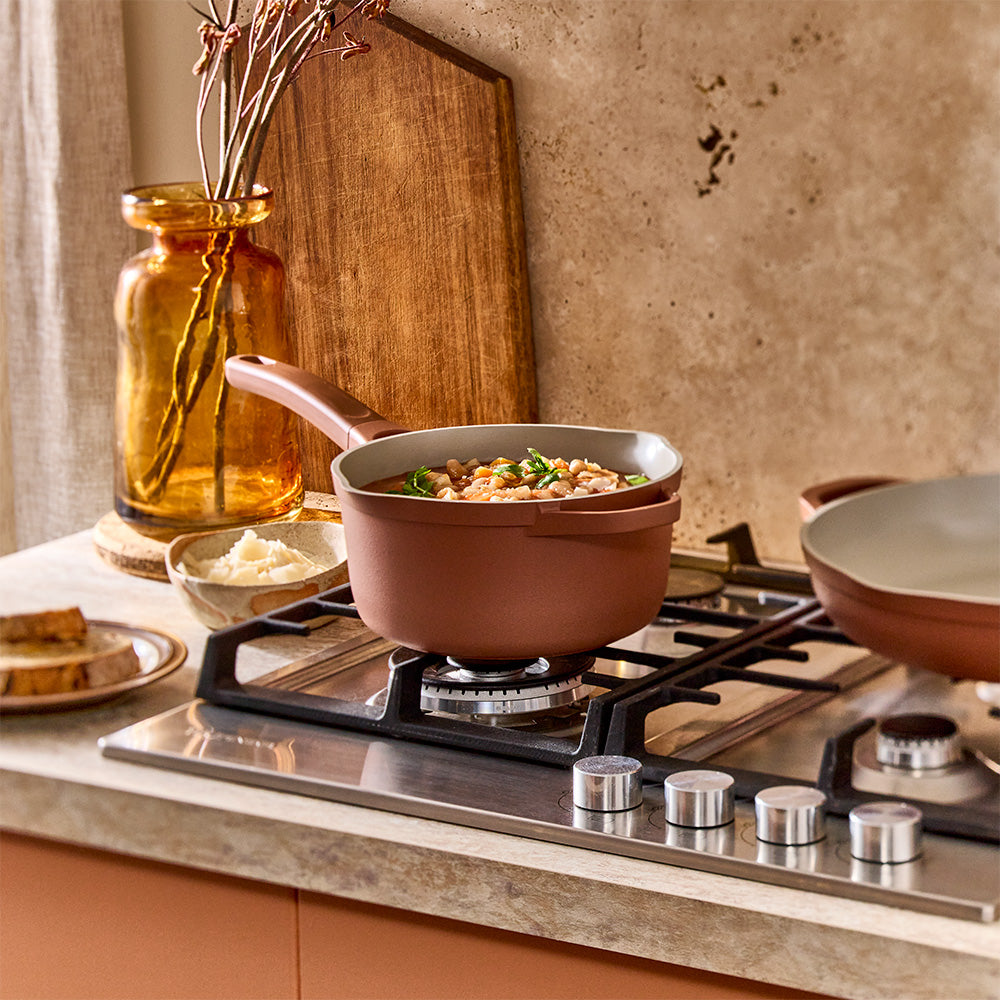 A terracotta-colored saucepan filled with soup sits on a cooktop burner in a kitchen setup.