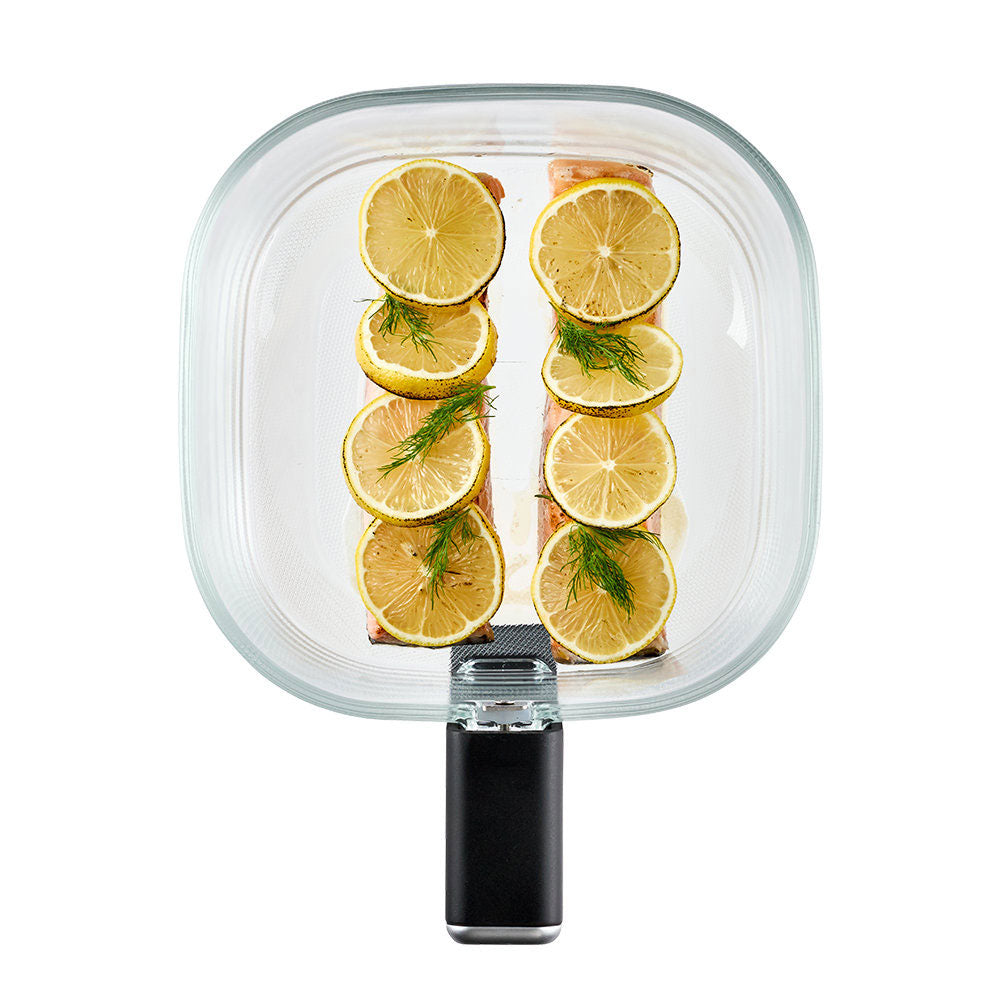 A clear glass air fryer with a black handle, shown from a top-down view containing two stacks of salmon fillets topped with lemon slices and dill.