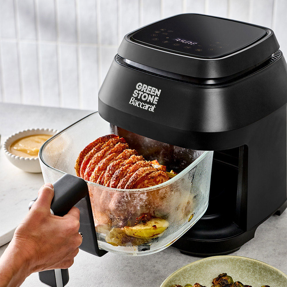 A hand is pulling the clear glass basket of a Green Stone Baccarat air fryer, which is shown containing cooked food. The air fryer is black with a digital control panel on top, set on a kitchen countertop.