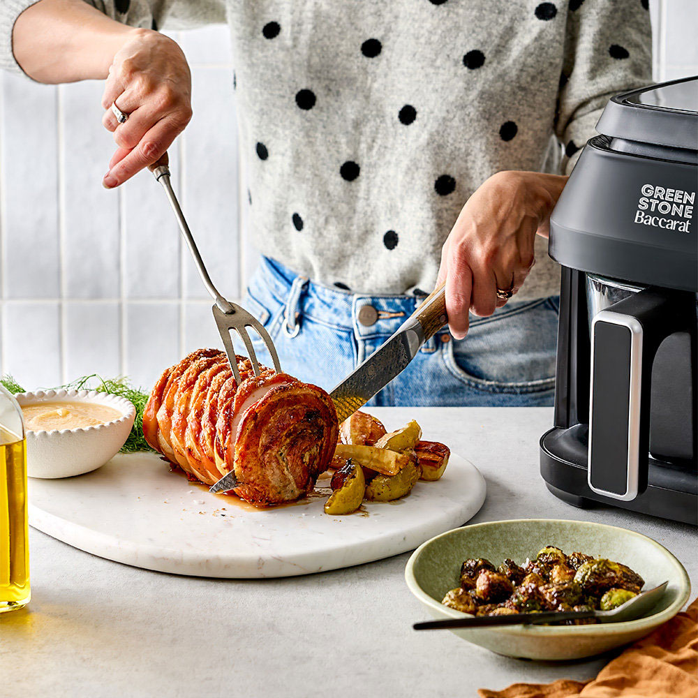A person slices a roasted meat on a white marble board with a fork and knife, with a countertop coffee maker labeled Green Stone Bullecart visible on the right. Sides include potato wedges, a small bowl of sauce, and another bowl on a light countertop.