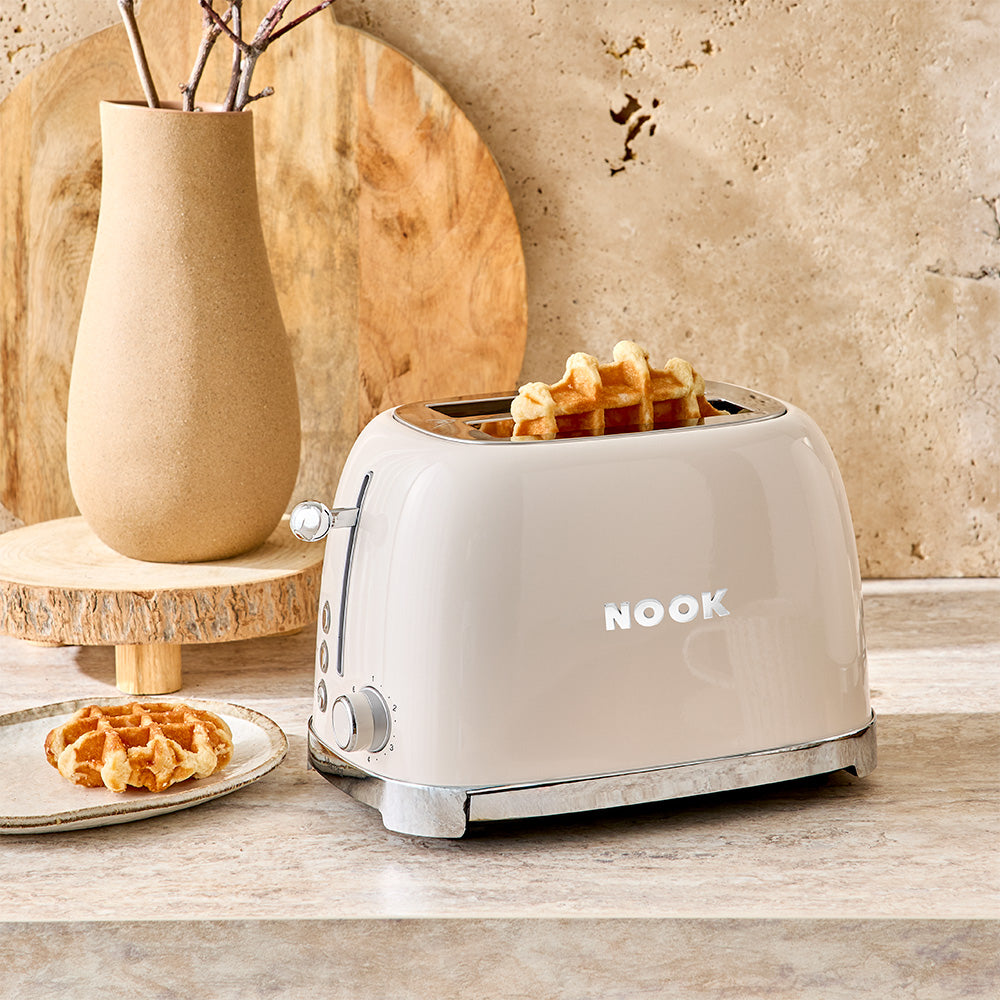 The cream toaster is positioned on a light-colored countertop in a styled kitchen, next to a wooden vase and a pastry, with a marble backsplash serving as the background.