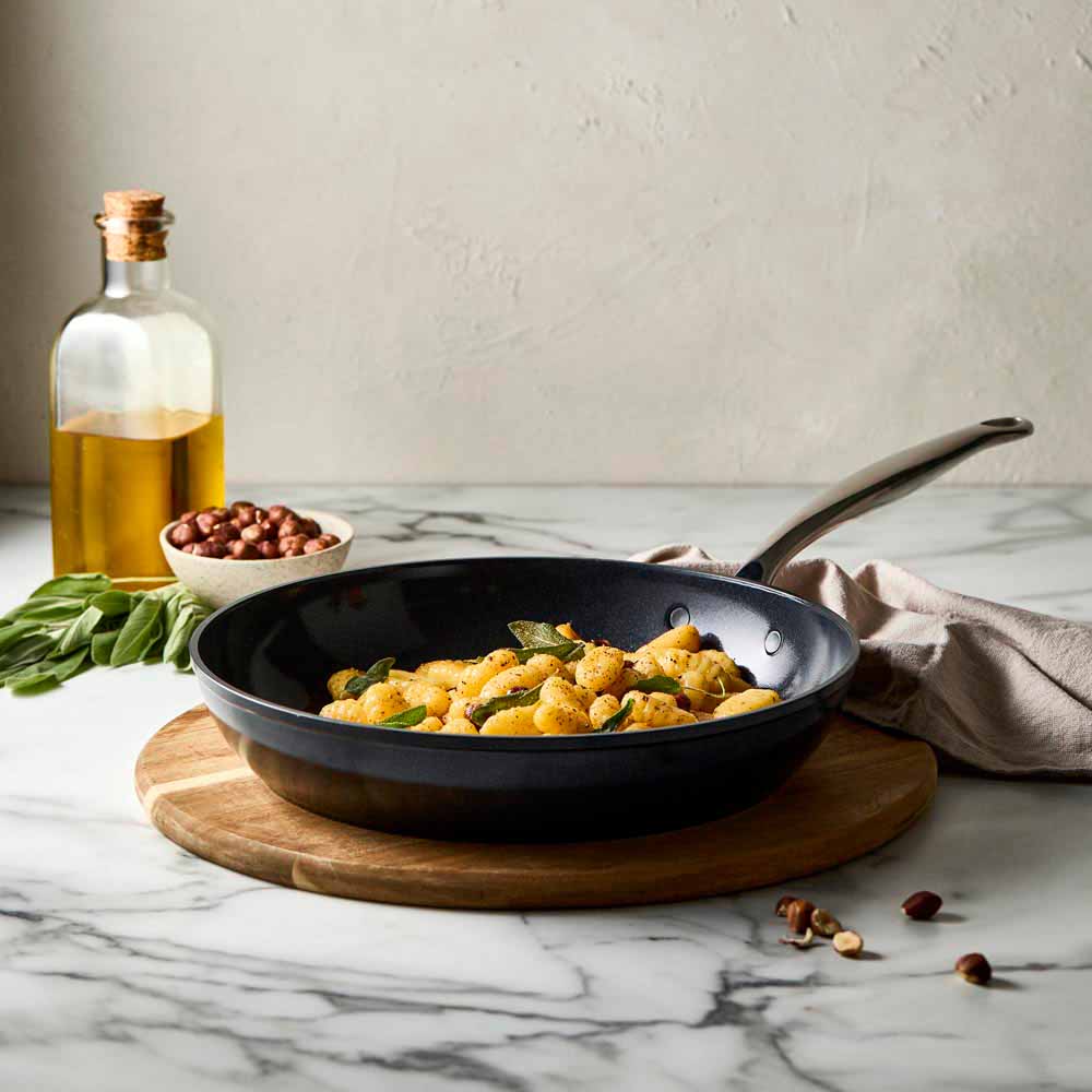 A Baccarat Le Connoisseurfrypan with pasta on a wooden board surrounded by olive oil and herbs
