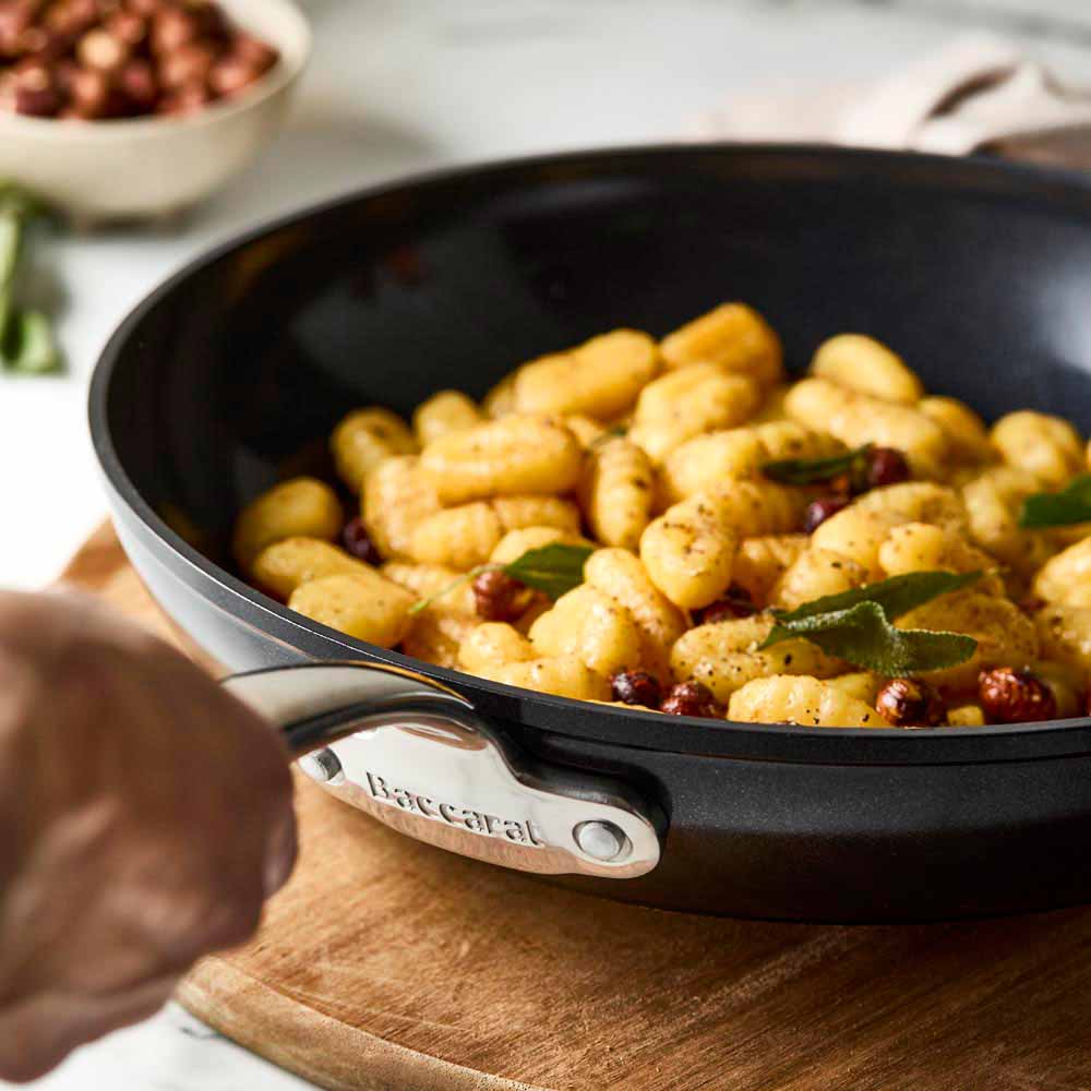 A Baccarat Le Connoisseurfrypan with pasta on a wooden board surrounded by olive oil and herbs