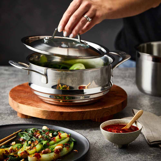 Hand lifting lid off stainless steel steamer with green vegetables on wooden board 32cm - Baccarat