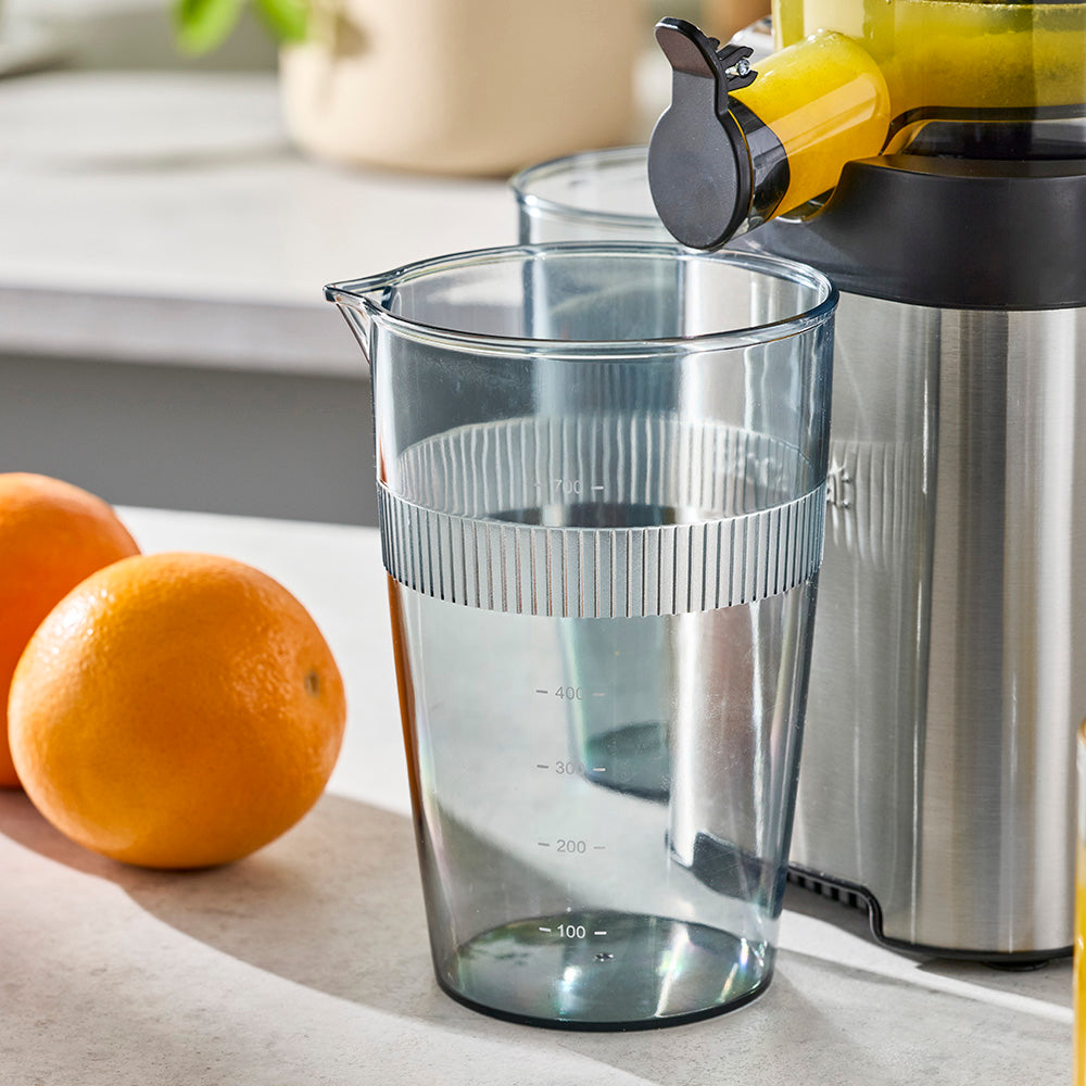 Clear juicer pitcher with a black lid on a kitchen counter with oranges in the background