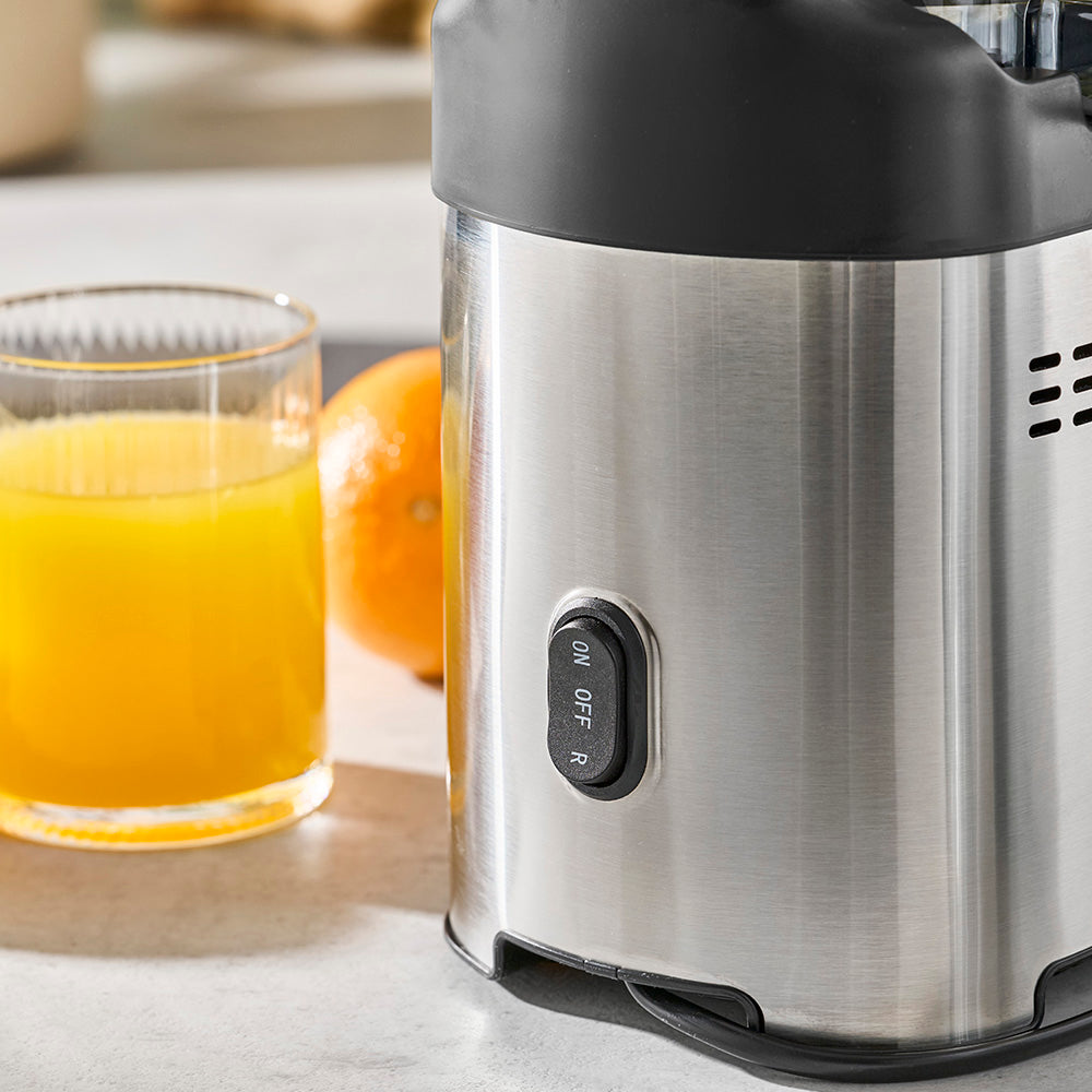 Stainless steel juicer with a glass of orange juice and an orange on a marble surface
